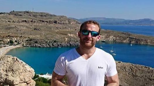 A man in a white t-shirt and sunglasses is smiling at the camera. In the background there are mountains and a body of blue water with sailboats. 