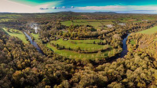A wide aerial view of a large park. The river Severn is curving through the middle of it, with thousands of trees in clusters on either side, as well as green fields. In the distance there are tall hills and white clouds.