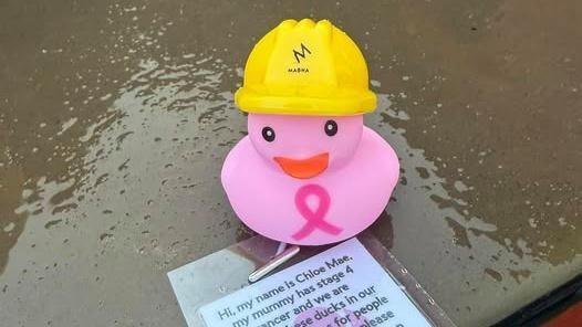 A pink rubber duck with a yellow hard hit on a wet slab of concrete. 