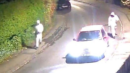 A CCTV image shows two men wearing white hazmat suits walking down a residential street with machetes. A car with headlights on is parked on the street.