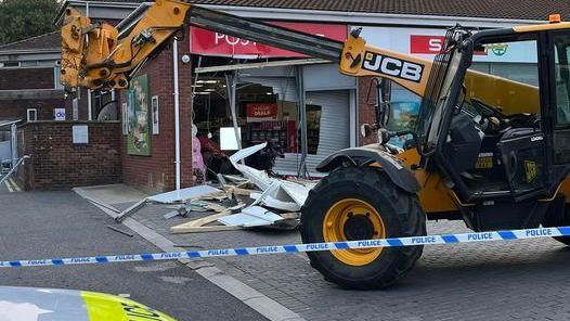 A yellow JCB digger in front of shop with a smashed up window and police tape around the scene 