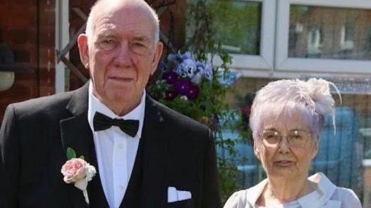 Roy and Susan Pollit pose for a photograph together. Roy wears a formal black suit with a black bow-tie and a pink flower in his button hole. Susan  wears a white fascinator and a light grey outfit.