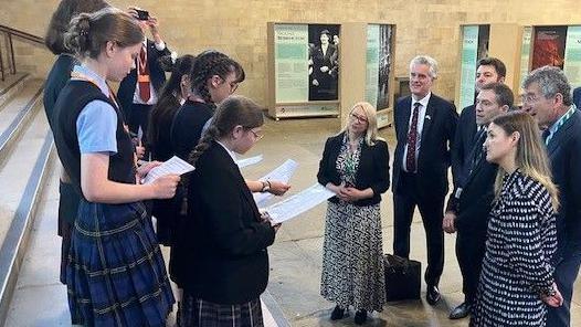 Six school students stand on steps inside Parliament. Six Mps are gathered in front listening.