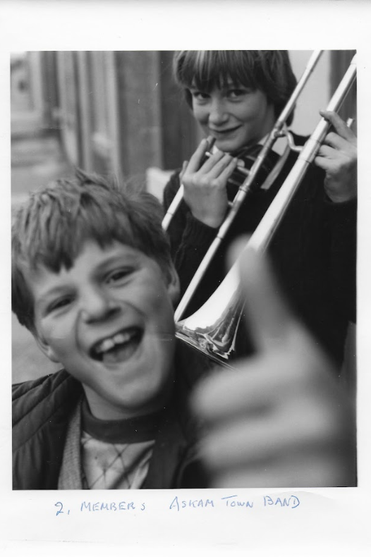 A black-and-white photo of two young boys looking into the camera. The one in the front has short, hair and is smiling with his mouth open, holding up his thumb which is out of focus. The boy in the back is smiling into the camera and has slightly longer hair. He is holding a trumpet and looks to be moving the instrument to his mouth. They are standing on a street with terraced housing.