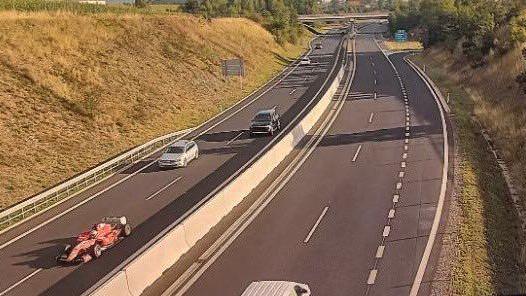 A red racing car speeds down the left had side of a motorway, flanked by normal cars.