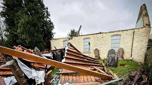 A stone church chapel, with it roof completely blown off. A pile of wood boards and beams covers the graveyard around the building