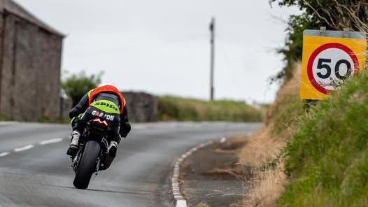 A rider in black with an orange bib negotiates a bend. There is a 50mph road sign above the grass bank on the right.