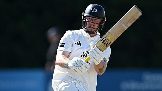 Miles Hammond pulls the ball away through mid-wicket. He is holding his bat high to the right of the picture. He is wearing his whites and has a dak blue helmet on