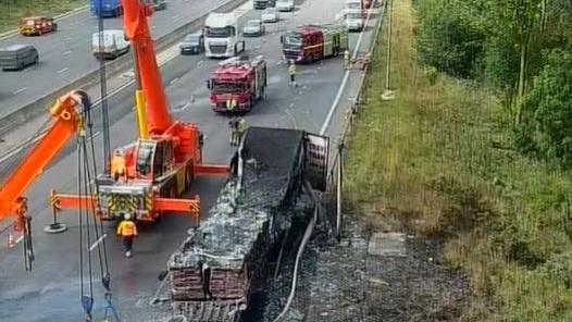 A blackened lorry being lifted by a recovery truck surrounded by fire engines