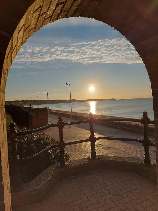 A photograph of the Bridlington seafront, taken behind a set of railings which are in shadow. The sun is rising. The sky is blue with hints of yellow and the sea is still and very blue. 