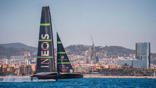 The Ineos Britannia yacht, with two triangular black and green sails and a black hull, at sea with Barcelona behind.