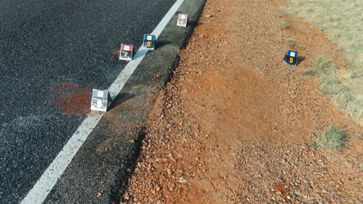 The side of a road, with red dirt and sparse grass. On the road and the dirt numbered markers are spaced out.