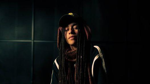 Bristol music artist Grove looks at the camera in a promotional photograph. They have long dreadlocks and is wearing a dark top and dark cap. The photograph is taken inside a dark room