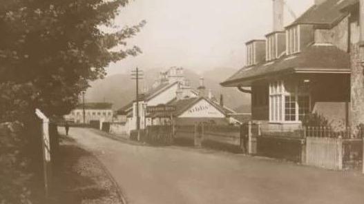 A black and white picture of the main street in Sandbank. There are more buildings in this picture than above and the hotel is far off in the distance. 