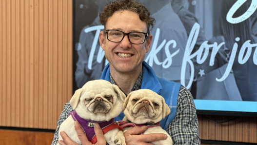 Mr Elshaw is holding two pugs and smiling at the camera. He is stood in front of a blue sign and is wearing a checked shirt and blue sleeveless top and has glasses on.