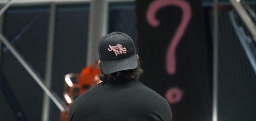 The back of a man with shoulder length black hair, wearing a black top and a black baseball hat, worn backwards with pink writing saying Jossiepops on it.
He appears to be looking a painting, covered in a black covering with a pink question mark on it.