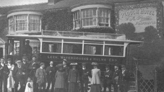 A black and white image shows people queuing outside the Plough Hotel for a bus. The building can be seen in the background with two large bay windows on the ground and first floors, and a mural with the name of the pub.