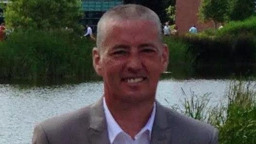 A man in a smart grey suit and white shirt, with short grey hair, smiles at the camera. Behind him is a large pond and greenery.