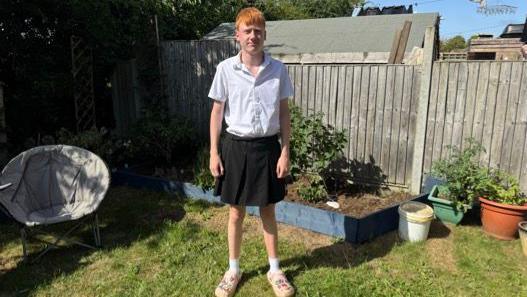 George is wearing a white short-sleeved shirt tucked into a dark pleated skirt, along with white socks and light-colored slip-on croc shoes. He is standing on a grassy lawn in a back garden facing forward with arms relaxed at his sides. Behind him is a wooden fence with raised garden beds containing soil and plants, along with several potted plants and a bucket. To the left side of the image is a round outdoor light grey-colored chair.