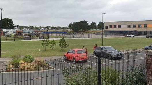 North Denes Primary School and Nursery, pictured from outside the black school fence. There is a car park in the foreground with a red Micra and a grey saloon parked, and a person getting into both cars. Behind that is a field and the school buildings and playground are in the background. It looks like a recently build school.