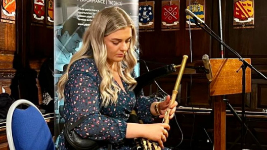 A young woman with blond long wavy hair playing the uilleann pipes on a blue chair. Behind her are brown panels with emblems and a grey pop-up sign. She is looking down at the uilleann pipes.