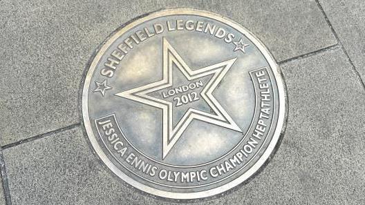 A gold-bronze coloured, circular metal plaque on the paving stone floor. Around the edge it reads "Sheffield Legends - Jessica Ennis Olympic champion heptathlete". In the centre is a star, with "London 2012" written in the middle of it.