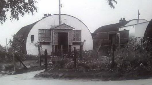 A Nissen hut with a porch added, the front painted white, and a fence raound it.