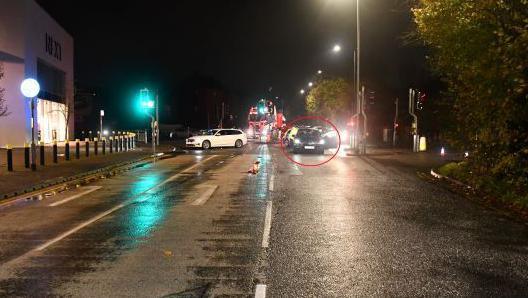 A view of the crossing where the collision happened, it is a dark, rainy evening and a black car can be seen in a red circle, highlighted as Roberts' car. Police cars and other vehicles can be seen in the background. 