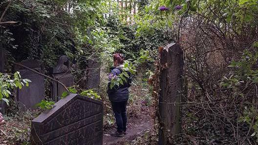 A woman stands in a graveyard looking at stones. There are a lot of trees and shrubs growing around Victorian grave stones.