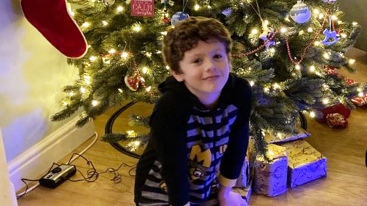 Dylan Cope who has curly brown hair and is wearing a black hoody with minion characters on it, sitting under a Christmas tree with presents next to him