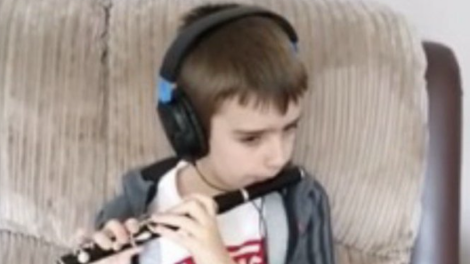 Shows a boy in a white and red t-shirt, grey and black hoodie and headphones, playing a black flute. He is sitting on a brown leather sofa.