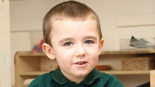 Lincoln Button is wearing a green school top. He is in a schoolroom, and behind him are some wooden shelves.