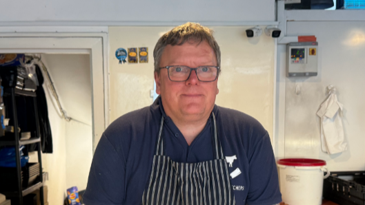 A man wearing a blue top and a striped apron stands inside a Butchers shop. He is smiling at the camera.