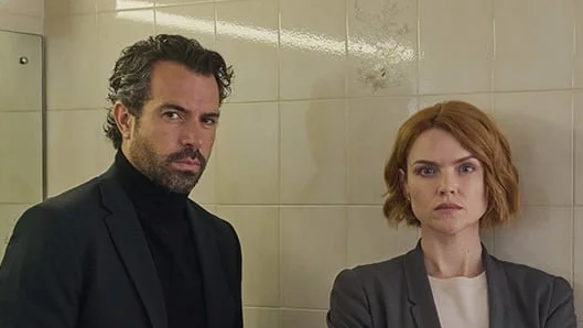 Tom Cullen and Erin Richards stood in a bathroom, with white tiles and mirrors behind them. Tom, on the left, is wearing a black suit and black polo neck top and has curly dark hair and a beard. Erin, on the right, has short curled ginger hair and is wearing a grey suit and white top. They are both looking at the camera with serious expressions.