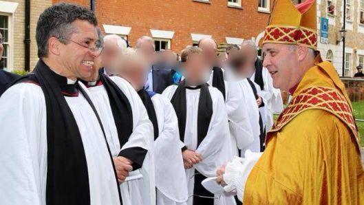 Former Anglican priest David Tudor, dressed in the standard black and white cassock, beams at Stephen Cottrell, then Bishop of Chelmsford, who is smiling back. Cottrell is dressed in golden robes and mitre worn by Anglican bishops. Behind them stand several other priests whose faces have been blurred to protect their identities.