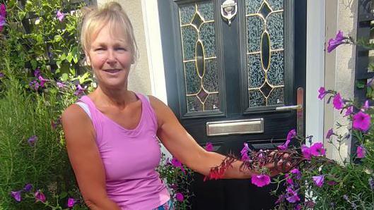 A woman with blonde hair and a full fringe tied back is wearing a bright pink vest and blue and pink shorts. She is smiling at the camera and standing next to a black and silver front door. Next to the front door are several purple flowers and greenery that she has planted.