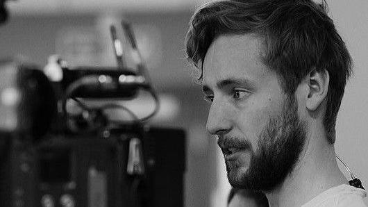 A black and white image of a man with a beard looking to the side. He has a white T-shirt on.