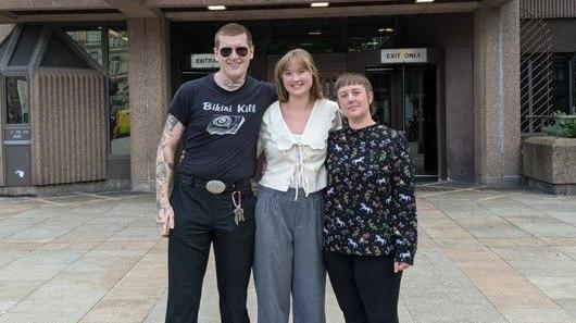 A man stands next to two woman outside Liverpool Crown Court. They have their arms around each other and are smiling. 