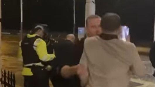 Colin Allan, in a cream blazer, holds a phone in his right hand while an officer grabs him