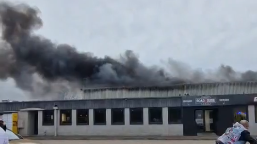 Black smoke pours out of the roof of an industrial building. 