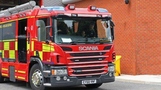 The front of a fire engine which is red and has fluorescent yellow squares down the side. Inside the engine on person driving can been seen and another person is sitting in the passenger seat.