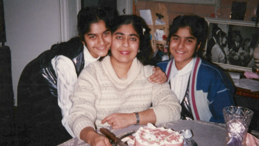 A family photograph of Annsa Chaudhry, wearing a white jumper and sitting at a table with a birthday cake on it, with her daughters either side of her.