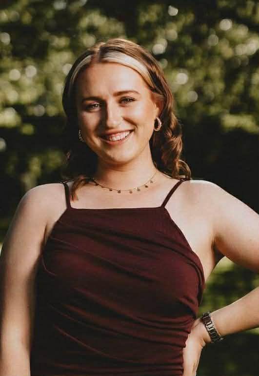 Një grua me flokë të kuq të çelët dhe nuanca bjonde i buzëqesh kamerës. Ajo ka veshur një fustan të kuq, një varëse dhe vathë me unaza. Ajo qëndron në një kopsht me gjethe të gjelbra pas saj.