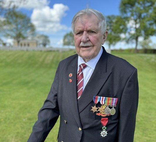 Arthur Oborne a world war two veteran stands on the grass at a military cemetery. He wears a blue blazer and army medals