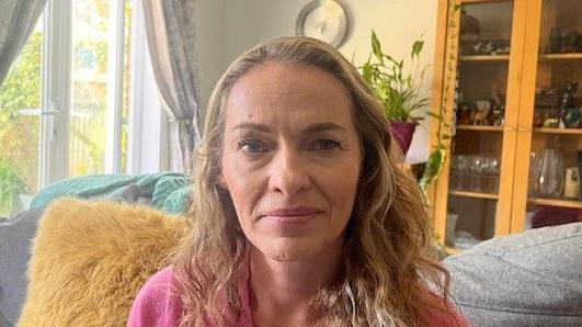 A woman with shoulder length blonde hair is looking at the camera. She is wearing a pink top. Behind her is a grey sofa with a beige cushion, a window, and a cupboard with glasses and vases visible inside