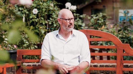 man with glasses sits on a bench surrounded by shrubbery