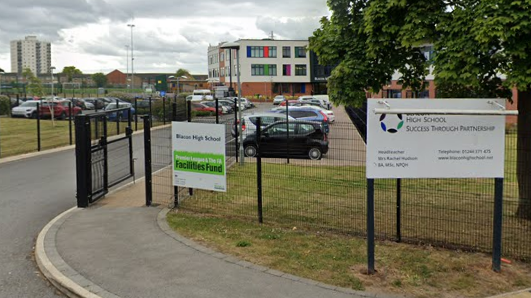 The exterior of Blacon High School shows two signs and a staff car park. In the background there is a brick two-storey building and to the left and right there are grass fields.