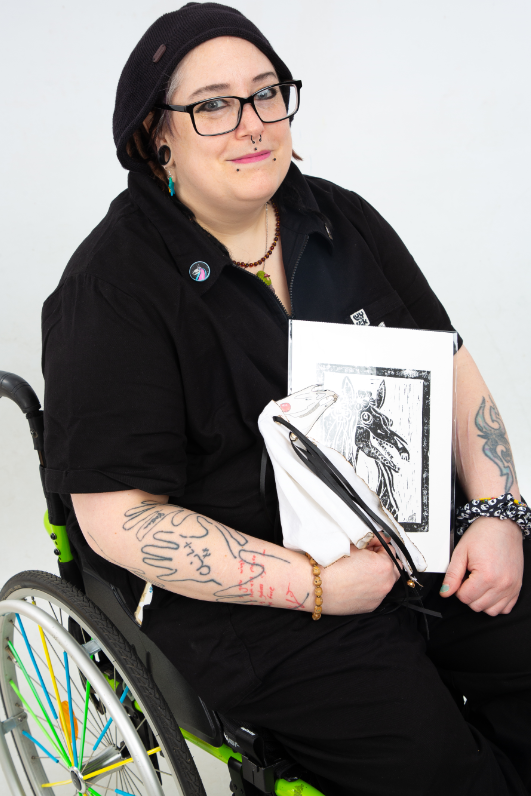 Woman with a number of face piercings has black hair. She's holding her artwork and is sitting in a wheelchair.
