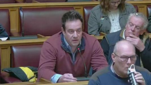 A man with short dark hair wearing a shirt and burgundy sweater sits in a council chamber. He is speaking into a microphone. There are two other men and a woman sitting on the wooden benches. 