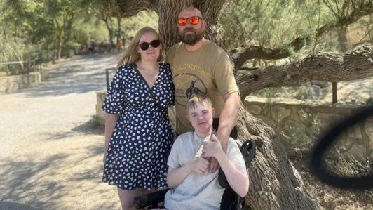 Michele, a woman with golden hair, wearing a navy blue and white dots knee-length dress, standing next to her husband , a man in a brown T-shirt, wearing orange sunglasses and their son, Oliver, sitting in a wheelchair in front of them. They are standing in front of a large tree.